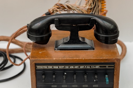 An antique rotary-style telephone rests on a wooden base with visible cords and a series of labeled switches, possibly indicating a multi-line or switchboard functionality.