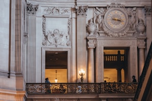 An ornate interior with classical architectural details such as intricately carved moldings and columns. A large clock with Roman numerals is centered above two large windows. Chandeliers cast a warm glow, and there is a metal railing with gold accents.