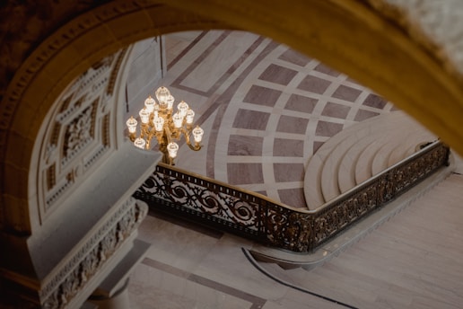 A grand interior space featuring an ornate chandelier with glowing lights hanging over a checkered marble floor. The scene is viewed through an archway with intricate carvings, leading to a beautiful staircase with wrought iron railings. The elegant architecture exhibits classical design elements.