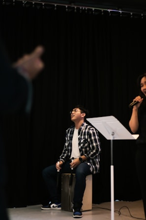 A person in a checkered shirt and jeans is sitting on a cajón, playing music. Another person is partially visible, holding a microphone. The setting appears to be a stage or performance area with black curtains in the background and a music stand nearby.