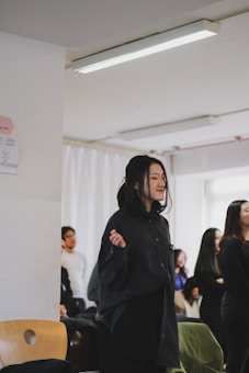 A group of people, with a focus on a young woman standing near a wall with her eyes closed, possibly deep in thought or prayer. The setting appears to be an indoor space with white walls and a fluorescent ceiling light. There are other individuals in the background, some of whom are seated and others standing.