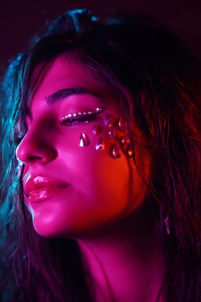 A close-up profile of a person's face adorned with rhinestones around the eyes, highlighted by dramatic pink and blue lighting. The expression is serene, with a focus on the intricate design of gems and the glossy appearance of the skin.