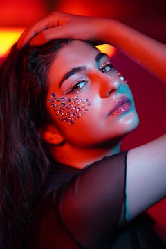 A close-up portrait of an individual with sparkling makeup and gemstones adorning the cheek area. The lighting casts a vibrant and dramatic effect, blending warm reds and cool blues for an artistic look. The hand is gently placed on the head, adding a dynamic pose to the composition.
