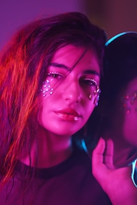 A person with long, flowing hair adorned with facial jewels and makeup is captured in vibrant lighting. The colors create a soft, glowing effect across the image, with the person holding a reflective object that adds a futuristic feel.