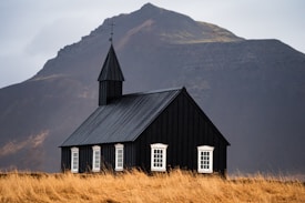 A small black church with white-trimmed windows stands prominently in a grassy field, framed by a rugged mountain in the background under an overcast sky. The structure is simple, featuring a pointed steeple topped with a cross.