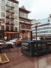 The image depicts an urban street scene featuring a mix of architectural styles. A traditional building with Asian architectural elements stands next to a more modern structure with glass windows. A subway entrance is in the foreground, marked with signage for Canal Street Station, which serves Uptown and The Bronx. Several vehicles, including a yellow taxi, are visible on the wet street, reflecting overcast or rainy weather conditions.