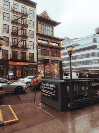 The image depicts an urban street scene featuring a mix of architectural styles. A traditional building with Asian architectural elements stands next to a more modern structure with glass windows. A subway entrance is in the foreground, marked with signage for Canal Street Station, which serves Uptown and The Bronx. Several vehicles, including a yellow taxi, are visible on the wet street, reflecting overcast or rainy weather conditions.