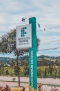 A tall, rectangular sign stands prominently, displaying the text 'INSTITUTO FEDERAL Minas Gerais' and 'CAMPUS RIBEIRÃO DAS NEVES.' The sign is positioned alongside a paved walkway with neatly maintained landscaping, including small trees and shrubs. In the background, there are distant hills and a cloudy sky.