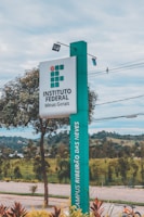 A tall, rectangular sign stands prominently, displaying the text 'INSTITUTO FEDERAL Minas Gerais' and 'CAMPUS RIBEIRÃO DAS NEVES.' The sign is positioned alongside a paved walkway with neatly maintained landscaping, including small trees and shrubs. In the background, there are distant hills and a cloudy sky.
