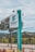 A tall, rectangular sign stands prominently, displaying the text 'INSTITUTO FEDERAL Minas Gerais' and 'CAMPUS RIBEIRÃO DAS NEVES.' The sign is positioned alongside a paved walkway with neatly maintained landscaping, including small trees and shrubs. In the background, there are distant hills and a cloudy sky.