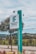 A tall, rectangular sign stands prominently, displaying the text 'INSTITUTO FEDERAL Minas Gerais' and 'CAMPUS RIBEIRÃO DAS NEVES.' The sign is positioned alongside a paved walkway with neatly maintained landscaping, including small trees and shrubs. In the background, there are distant hills and a cloudy sky.