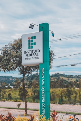 A tall, rectangular sign stands prominently, displaying the text 'INSTITUTO FEDERAL Minas Gerais' and 'CAMPUS RIBEIRÃO DAS NEVES.' The sign is positioned alongside a paved walkway with neatly maintained landscaping, including small trees and shrubs. In the background, there are distant hills and a cloudy sky.