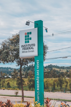 A tall, rectangular sign stands prominently, displaying the text 'INSTITUTO FEDERAL Minas Gerais' and 'CAMPUS RIBEIR&Atilde;O DAS NEVES.' The sign is positioned alongside a paved walkway with neatly maintained landscaping, including small trees and shrubs. In the background, there are distant hills and a cloudy sky.
