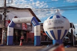An urban scene featuring an inflatable archway and balloon with the text 'Ribeirão das Neves Prefeitura'. The structures are set up in front of a small store with a red and beige exterior. A person with a backpack is walking through the archway, and there's a traffic cone and electrical wires overhead.