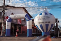 An urban scene featuring an inflatable archway and balloon with the text 'Ribeir&atilde;o das Neves Prefeitura'. The structures are set up in front of a small store with a red and beige exterior. A person with a backpack is walking through the archway, and there's a traffic cone and electrical wires overhead.