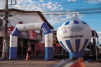 An urban scene featuring an inflatable archway and balloon with the text 'Ribeirão das Neves Prefeitura'. The structures are set up in front of a small store with a red and beige exterior. A person with a backpack is walking through the archway, and there's a traffic cone and electrical wires overhead.