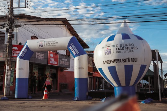 An urban scene featuring an inflatable archway and balloon with the text 'Ribeir&atilde;o das Neves Prefeitura'. The structures are set up in front of a small store with a red and beige exterior. A person with a backpack is walking through the archway, and there's a traffic cone and electrical wires overhead.