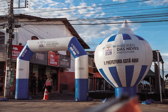 An urban scene featuring an inflatable archway and balloon with the text 'Ribeir&atilde;o das Neves Prefeitura'. The structures are set up in front of a small store with a red and beige exterior. A person with a backpack is walking through the archway, and there's a traffic cone and electrical wires overhead.