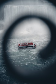 A tour boat filled with people in red rain ponchos navigates through choppy waters. The boat is positioned against the backdrop of a large, misty waterfall, partially obscured by a circular frame in the foreground, creating a dramatic perspective.