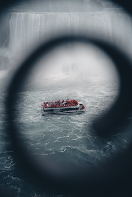 A tour boat filled with people in red rain ponchos navigates through choppy waters. The boat is positioned against the backdrop of a large, misty waterfall, partially obscured by a circular frame in the foreground, creating a dramatic perspective.