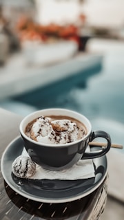 A black cup filled with a creamy beverage topped with whipped cream and chocolate powder sits on a matching saucer. Next to the cup is a small, powdered pastry on a napkin, accompanied by a stirrer. The background features a blurred outdoor setting with hints of a pool and foliage.