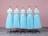 Five dress forms are aligned in a row, each displaying an identical light blue, sleeveless dress with a fitted bodice and a full skirt. They are placed on a gray patterned floor in front of a soft pink background.