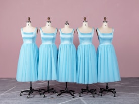 Five dress forms are aligned in a row, each displaying an identical light blue, sleeveless dress with a fitted bodice and a full skirt. They are placed on a gray patterned floor in front of a soft pink background.