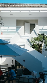 A modern outdoor patio with white staircases leading up to an open door on the second level. The sunny setting includes a large white umbrella providing shade over a small seating area with a person dressed in blue sitting alone. A black guitar rests against a wall, and there's a potted plant near the stairs. The sky is bright blue overhead.