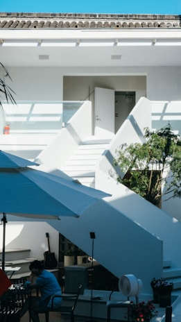 A modern outdoor patio with white staircases leading up to an open door on the second level. The sunny setting includes a large white umbrella providing shade over a small seating area with a person dressed in blue sitting alone. A black guitar rests against a wall, and there's a potted plant near the stairs. The sky is bright blue overhead.