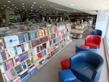 A spacious bookstore with well-organized shelves filled with a variety of books. The shelves are labeled with categories like science, fiction, and classics. Comfortable red and blue chairs are arranged along the window, providing a cozy reading area. Large windows allow natural light to flood the space, creating a warm atmosphere.