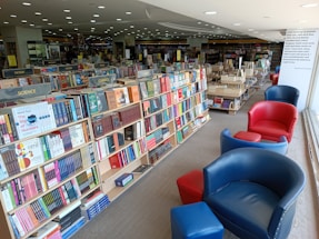 A spacious bookstore with well-organized shelves filled with a variety of books. The shelves are labeled with categories like science, fiction, and classics. Comfortable red and blue chairs are arranged along the window, providing a cozy reading area. Large windows allow natural light to flood the space, creating a warm atmosphere.