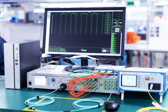 A laboratory setup featuring various networking and testing equipment on a table. A computer monitor displaying test results with a 'Pass' message is prominently positioned. Multiple cables in orange and blue are connected to devices labeled 'Optical Phase Modulation Meter' and 'Test Station'. The environment appears clean and organized, suggesting a professional or industrial setting.