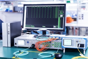 A laboratory setup featuring various networking and testing equipment on a table. A computer monitor displaying test results with a 'Pass' message is prominently positioned. Multiple cables in orange and blue are connected to devices labeled 'Optical Phase Modulation Meter' and 'Test Station'. The environment appears clean and organized, suggesting a professional or industrial setting.