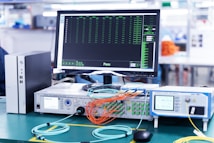 A laboratory setup featuring various networking and testing equipment on a table. A computer monitor displaying test results with a 'Pass' message is prominently positioned. Multiple cables in orange and blue are connected to devices labeled 'Optical Phase Modulation Meter' and 'Test Station'. The environment appears clean and organized, suggesting a professional or industrial setting.