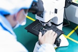 A person in laboratory attire closely examines a specimen through a microscope on a green work surface. The individual is adjusting the stage of the microscope and wearing protective clothing.