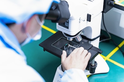 A person in laboratory attire closely examines a specimen through a microscope on a green work surface. The individual is adjusting the stage of the microscope and wearing protective clothing.