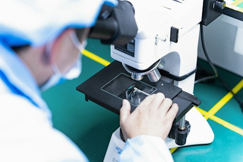 A person in laboratory attire closely examines a specimen through a microscope on a green work surface. The individual is adjusting the stage of the microscope and wearing protective clothing.
