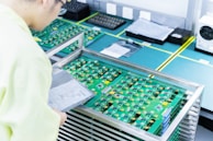 A person wearing light-colored clothing examines a stack of green circuit boards placed in a metal rack on a workbench. The boards have various components attached and are surrounded by other technological equipment in a clean and organized workspace.