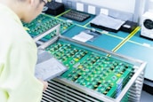 A person wearing light-colored clothing examines a stack of green circuit boards placed in a metal rack on a workbench. The boards have various components attached and are surrounded by other technological equipment in a clean and organized workspace.