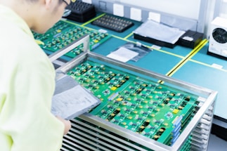 A person wearing light-colored clothing examines a stack of green circuit boards placed in a metal rack on a workbench. The boards have various components attached and are surrounded by other technological equipment in a clean and organized workspace.