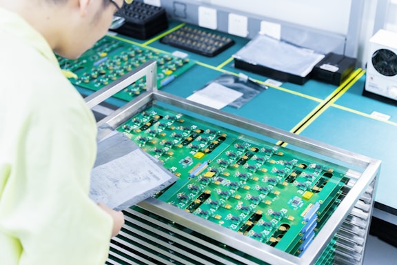A person wearing light-colored clothing examines a stack of green circuit boards placed in a metal rack on a workbench. The boards have various components attached and are surrounded by other technological equipment in a clean and organized workspace.