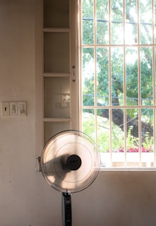 A sunlit room features a standing electric fan positioned in front of a large window with bars. Outside, a lush tree and blooming flowers in a planter box can be seen, partially obscured by the window. The soft light creates gentle shadows on the wall.