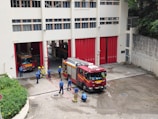 A group of firefighters are gathered outside a fire station. Two fire trucks are visible, one parked inside the station and another one parked outside. The firefighters are wearing blue uniforms and yellow helmets, and appear to be engaged in a discussion or training exercise. The building is off-white with red doors, and there are some green plants along the side.