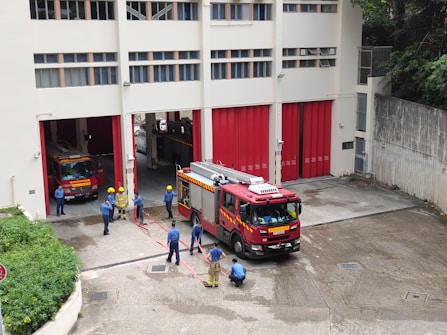 A group of firefighters are gathered outside a fire station. Two fire trucks are visible, one parked inside the station and another one parked outside. The firefighters are wearing blue uniforms and yellow helmets, and appear to be engaged in a discussion or training exercise. The building is off-white with red doors, and there are some green plants along the side.
