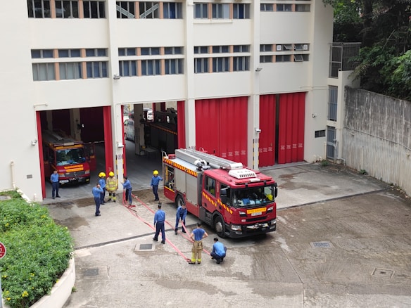 A group of firefighters are gathered outside a fire station. Two fire trucks are visible, one parked inside the station and another one parked outside. The firefighters are wearing blue uniforms and yellow helmets, and appear to be engaged in a discussion or training exercise. The building is off-white with red doors, and there are some green plants along the side.