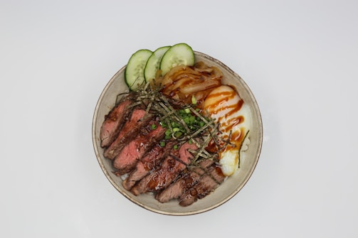 A bowl of food containing sliced medium-rare beef steak garnished with green onions and seaweed strips, slices of cucumber, caramelized onions, and a soft-boiled egg drizzled with a dark sauce.