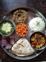 A traditional Indian meal served on a metal thali, featuring a variety of dishes. There is a portion of plain white rice, a serving of thick wheat-based porridge, and a flatbread. Accompaniments include a bowl of green curry, small crispy orange snacks, and a dish of colorful mixed vegetables. The meal is garnished with sliced red onions and fresh herbs.