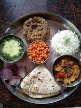 A traditional Indian meal served on a metal thali, featuring a variety of dishes. There is a portion of plain white rice, a serving of thick wheat-based porridge, and a flatbread. Accompaniments include a bowl of green curry, small crispy orange snacks, and a dish of colorful mixed vegetables. The meal is garnished with sliced red onions and fresh herbs.