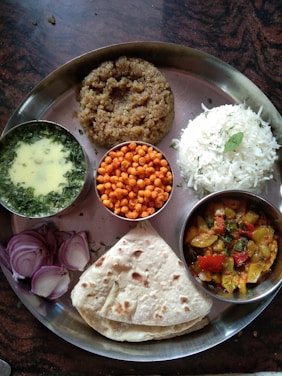 A traditional Indian meal served on a metal thali, featuring a variety of dishes. There is a portion of plain white rice, a serving of thick wheat-based porridge, and a flatbread. Accompaniments include a bowl of green curry, small crispy orange snacks, and a dish of colorful mixed vegetables. The meal is garnished with sliced red onions and fresh herbs.