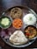 A traditional Indian meal served on a metal thali, featuring a variety of dishes. There is a portion of plain white rice, a serving of thick wheat-based porridge, and a flatbread. Accompaniments include a bowl of green curry, small crispy orange snacks, and a dish of colorful mixed vegetables. The meal is garnished with sliced red onions and fresh herbs.